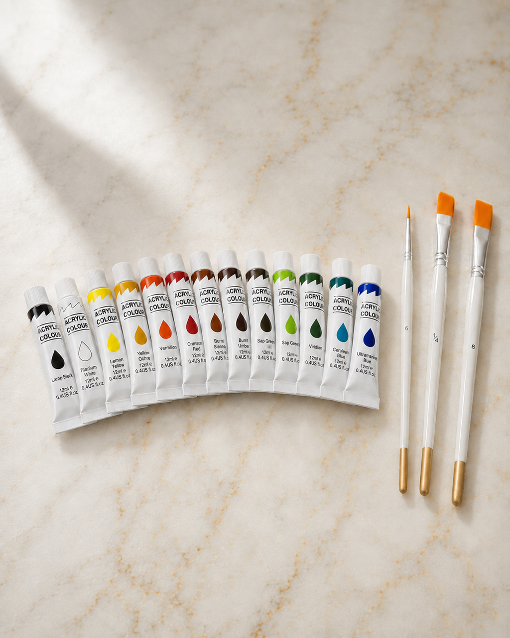 A set of 12 tubes of acrylic paint in various colors arranged in a semi-circle on a light beige marble surface, accompanied by three white paintbrushes with gold ferrules and orange tips.