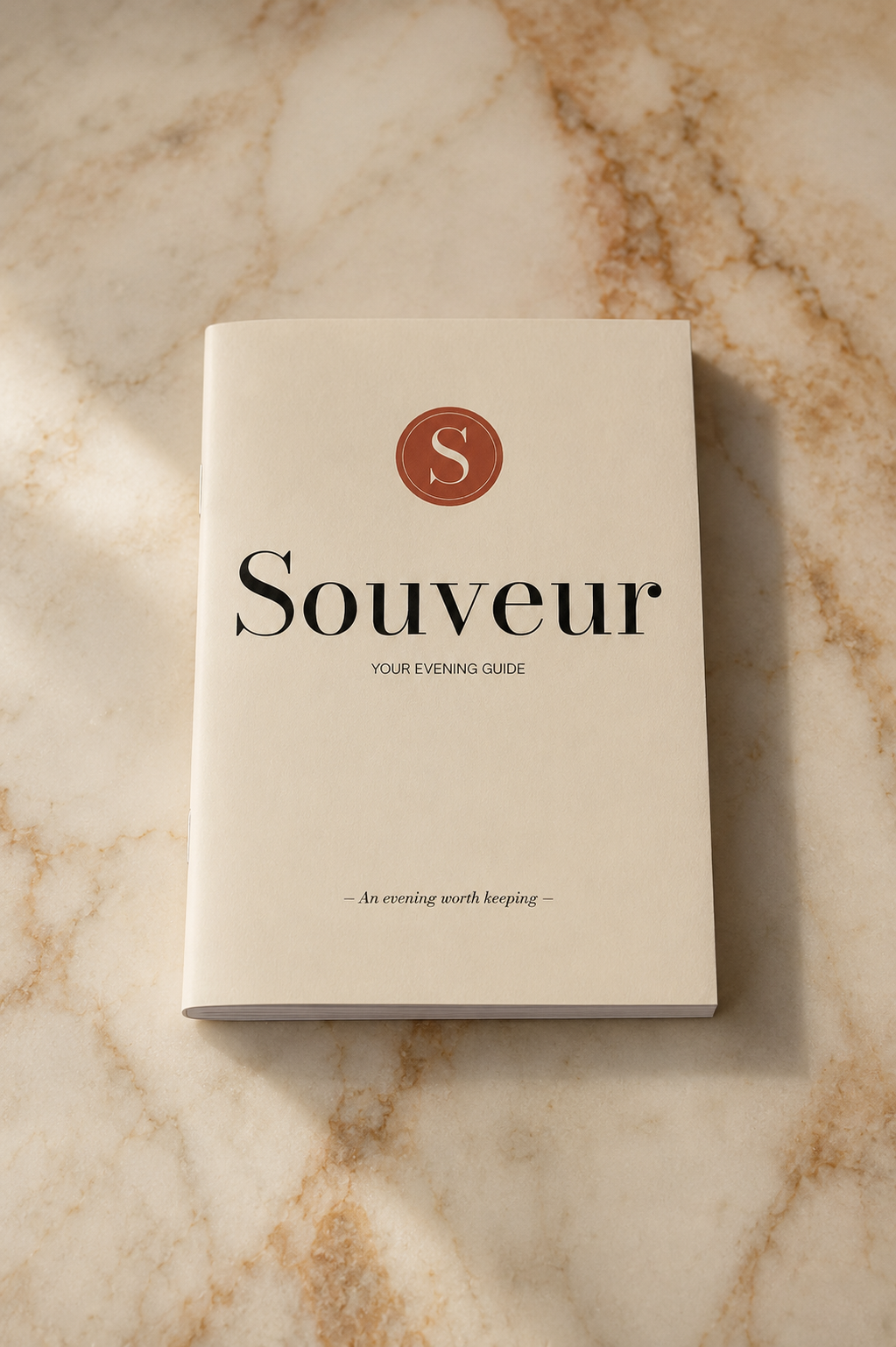 A square cream-colored book or guide titled 'Souvear' with a circular red 'S' logo at the top, placed on a beige marble surface with soft sunlight and shadows.