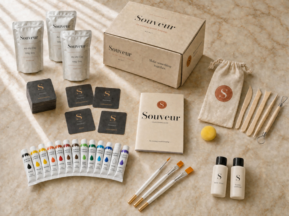 A Souveur DIY air-dry pottery and painting date-night kit arranged on a beige marble surface. The kit includes a kraft paper box, multiple pouches of air-dry clay, a small canvas pouch with a monogrammed 'S', wooden spatulas, a yellow mixing ball, two small bottles, several paint tubes, cards, and brushes.