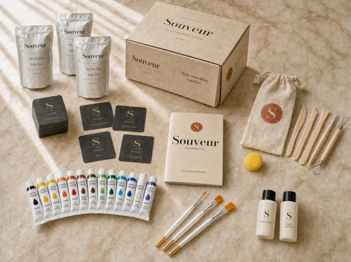 A Souveur DIY air-dry pottery and painting date-night kit arranged on a beige marble surface. The kit includes a kraft paper box, multiple pouches of air-dry clay, a small canvas pouch with a monogrammed 'S', wooden spatulas, a yellow mixing ball, two small bottles, several paint tubes, cards, and brushes.