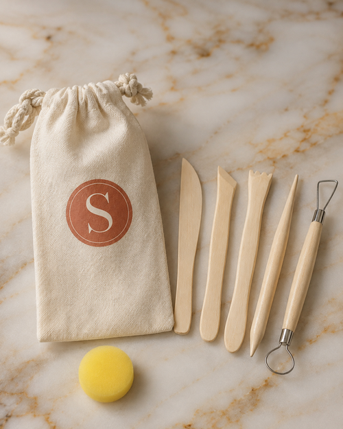 A clay sculpting kit with five wooden tools, a yellow sponge, and a cream drawstring pouch with a brown monogram 'S', arranged on a white marble background.